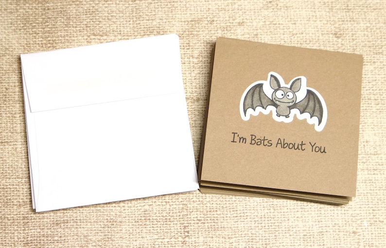 May include: A stack of brown cards with a cartoon bat illustration and the text "I'm Bats About You" next to a stack of white envelopes. The bat is gray with a white outline. The cards and envelopes are on a textured, tan surface.