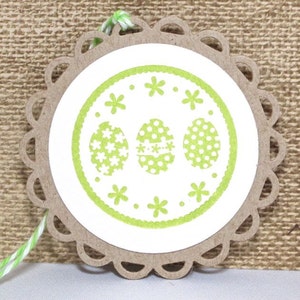 May include: A white paper circle with a green border and three green Easter eggs with white polka dots and white flowers. The circle is attached to a brown paper tag with a scalloped edge.
