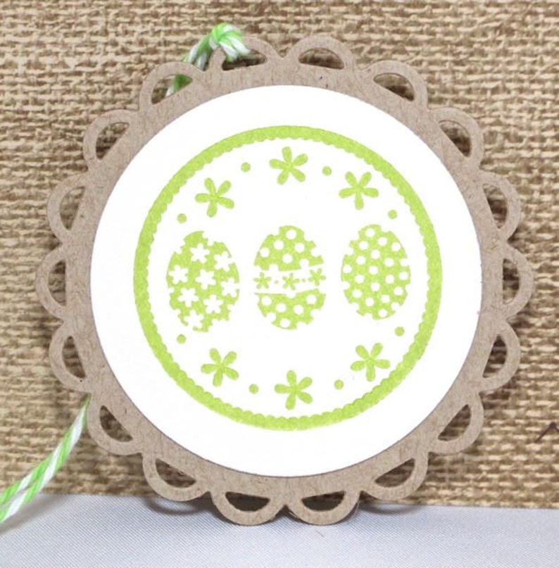 May include: A white paper circle with a green border and three green Easter eggs with white polka dots and white flowers. The circle is attached to a brown paper tag with a scalloped edge.
