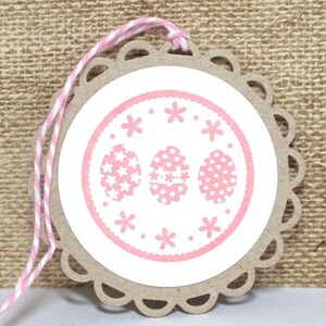 May include: A pink and white Easter gift tag with a scalloped edge and a pink circle with three pink eggs with white polka dots and white flowers. The tag is tied with a pink and white twine.