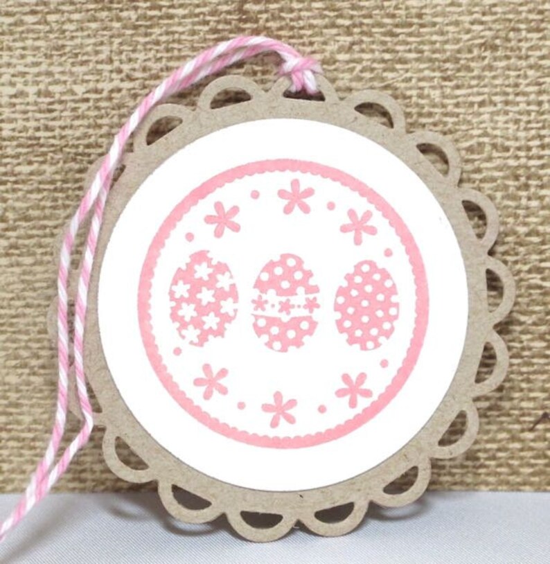May include: A pink and white Easter gift tag with a scalloped edge and a pink circle with three pink eggs with white polka dots and white flowers. The tag is tied with a pink and white twine.