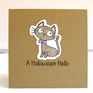 May include: A brown square card with a cartoon cat sticker. The gray cat has a purple bow tie and is waving. The text "A Halloween Hello" is printed below the cat.