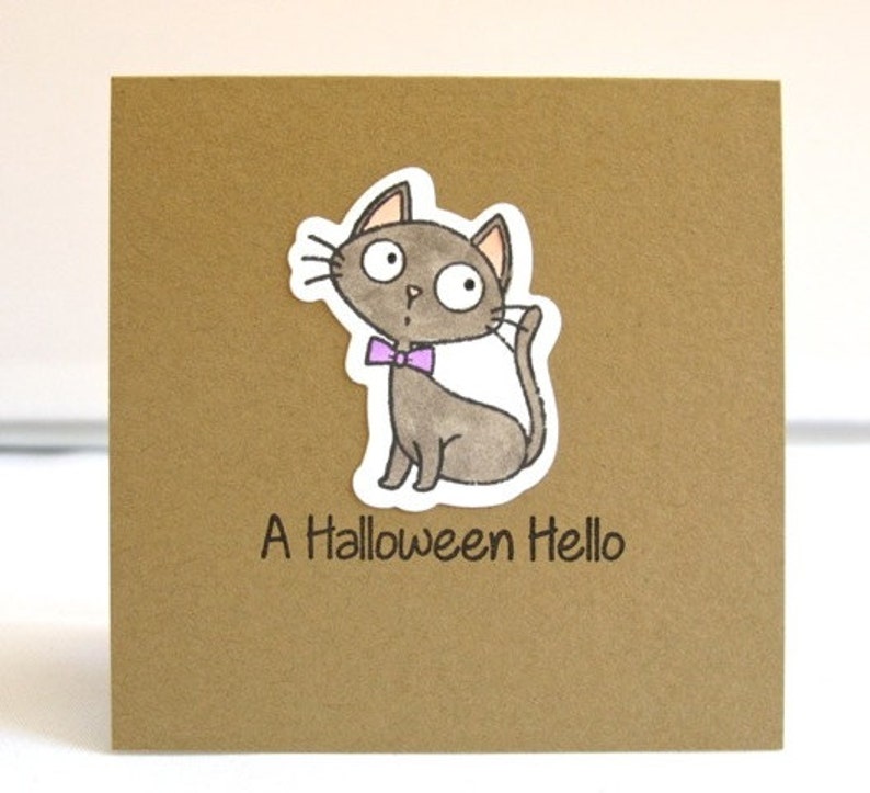 May include: A brown square card with a cartoon cat sticker. The gray cat has a purple bow tie and is waving. The text "A Halloween Hello" is printed below the cat.