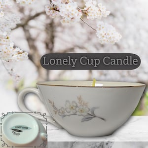 May include: A white teacup candle with a floral design and gold trim. The teacup has a handle and is filled with wax. The text "Lonely Cup Candle" is displayed on the image. The bottom of the teacup is also visible, with text that reads "Synco China Cerise 18757".