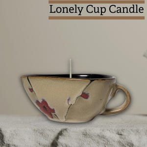 May include: A beige teacup candle with a black rim and a handle. The cup features red floral designs and the text "Lonely Cup Candle" at the top. A white candle wick is centered in the cup.