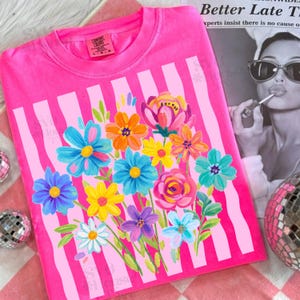 Op de afbeelding: Een felroze t-shirt met een grafische print van een kleurrijk boeket geschilderde bloemen in tinten blauw, geel, oranje, roze en paars. Het bloemmotief is geplaatst over een achtergrond van verticale lichtroze strepen op de stof.