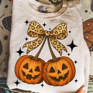 Puede incluir: Sudadera color crema con un diseño de Halloween. El diseño incluye dos calabazas naranjas con caras talladas, conectadas por tallos a un lazo con estampado de leopardo. Estrellas negras adornan las calabazas.