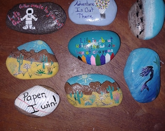 May include: Hand-painted rocks with various designs and inspirational quotes. One rock features an astronaut, another a hot air balloon with the text "Adventure Is Out There." Other rocks display desert landscapes, a mermaid, and the text "Paper, I win!"