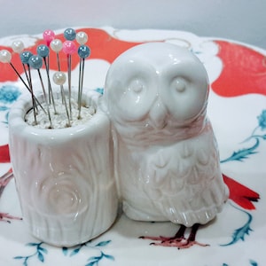 May include: White ceramic owl pincushion with a tree trunk pincushion next to it. The pincushion is filled with colorful pins.