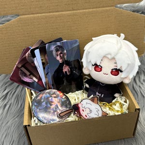 May include: A cardboard gift box containing a plush doll with white hair and red eyes, a circular pin featuring a character portrait, a small acrylic charm, and a set of rectangular trading cards displaying character illustrations in a dark color palette.