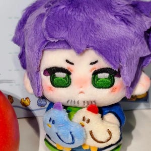 Könnte beinhalten: Eine Plüschpuppe mit lila Haaren, grünen Augen und einem blauen Hemd. Die Puppe hält zwei Plüschvögel, einen blauen und einen weißen. Die Puppe hat einen ernsten Gesichtsausdruck.