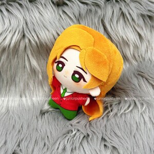 May include: A plush doll with a cartoon design. The doll has a yellow head and hair, green eyes, and is wearing a red jacket with a green tie and green trousers. The doll is on a grey, furry surface.