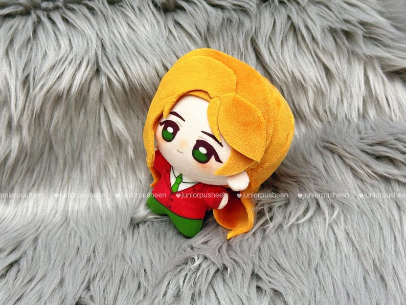 May include: A plush doll with a cartoon design. The doll has a yellow head and hair, green eyes, and is wearing a red jacket with a green tie and green trousers. The doll is on a grey, furry surface.