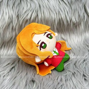 May include: A plush doll with a round head, large green eyes, and orange hair. The doll wears a red jacket with a green tie and green trousers. The doll is lying on a grey, furry surface.