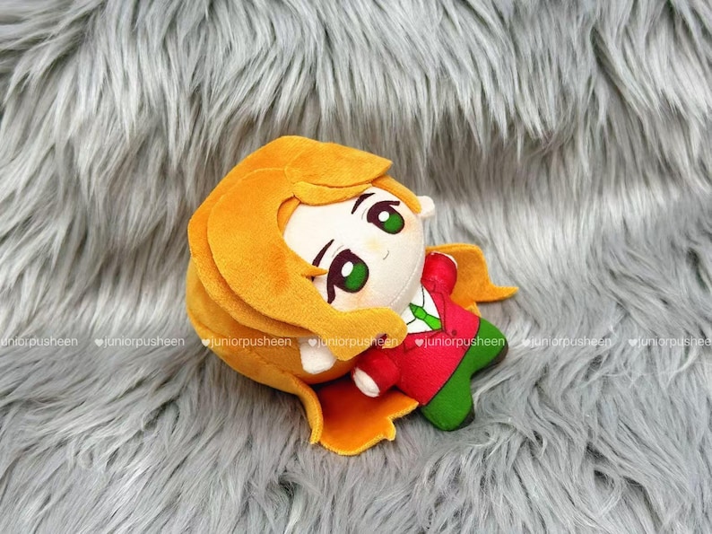 May include: A plush doll with a round head, large green eyes, and orange hair. The doll wears a red jacket with a green tie and green trousers. The doll is lying on a grey, furry surface.