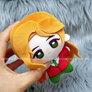 May include: A plush doll with a cartoon design. The doll has blonde hair, green eyes, and wears a red jacket, green trousers, and a green tie. The doll is held in a hand.