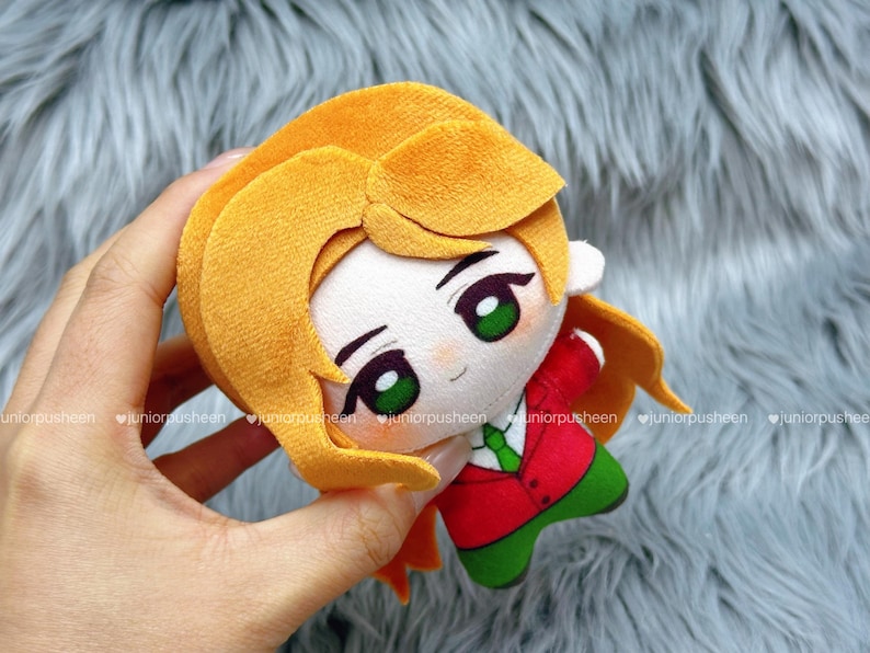 May include: A plush doll with a cartoon design. The doll has blonde hair, green eyes, and wears a red jacket, green trousers, and a green tie. The doll is held in a hand.