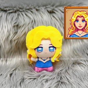 K&ouml;nnte beinhalten: Ein Pl&uuml;schpuppe eines Charakters mit gelben Haaren, blauen Augen und einem blau-rosa Outfit. Die Puppe befindet sich auf einer grauen, pelzigen Oberfl&auml;che. Ein gerahmtes Pixel-Art-Portr&auml;t des Charakters ist im Hintergrund.