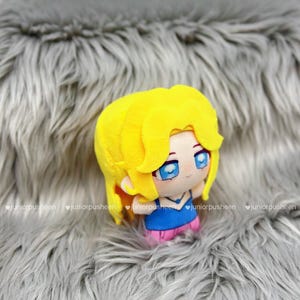 K&ouml;nnte beinhalten: Pl&uuml;schpuppe mit gelben Haaren, blauen Augen und einem blauen Oberteil mit rosa Shorts. Die Puppe ist vor einem grauen, pelzigen Hintergrund platziert. Das Design der Puppe erinnert an einen Anime-Charakter.