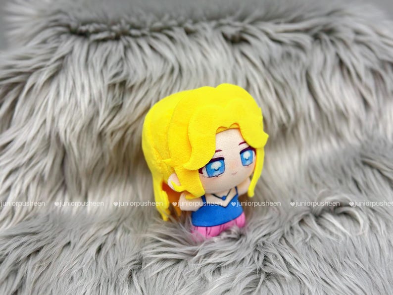 K&ouml;nnte beinhalten: Pl&uuml;schpuppe mit gelben Haaren, blauen Augen und einem blauen Oberteil mit rosa Shorts. Die Puppe ist vor einem grauen, pelzigen Hintergrund platziert. Das Design der Puppe erinnert an einen Anime-Charakter.