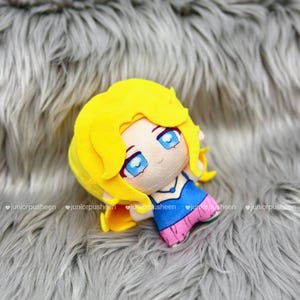 K&ouml;nnte beinhalten: Eine Pl&uuml;schpuppe mit gelben Haaren, blauen Augen und einem blau-rosa Outfit. Die Puppe liegt auf einer grauen, pelzigen Oberfl&auml;che. Das Gesicht der Puppe hat ein niedliches, cartoonartiges Design.