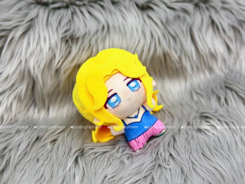 K&ouml;nnte beinhalten: Eine Pl&uuml;schpuppe mit gelben Haaren, blauen Augen und einem blau-rosa Outfit. Die Puppe liegt auf einer grauen, pelzigen Oberfl&auml;che. Das Gesicht der Puppe hat ein niedliches, cartoonartiges Design.