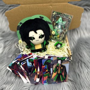 Puede incluir: Una caja de regalo que contiene un muñeco de peluche, cartas coleccionables y otros artículos. El muñeco de peluche tiene cabello negro y un atuendo verde y amarillo. Las cartas presentan ilustraciones coloridas.