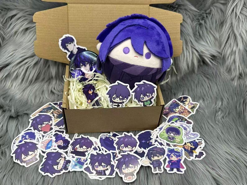Puede incluir: Una mu&ntilde;eca de peluche con cabello azul y una bufanda a juego se encuentra en una caja de cart&oacute;n llena de pegatinas y papel de relleno. Las pegatinas presentan varios dise&ntilde;os de personajes de estilo anime en tonos morados y azules.
