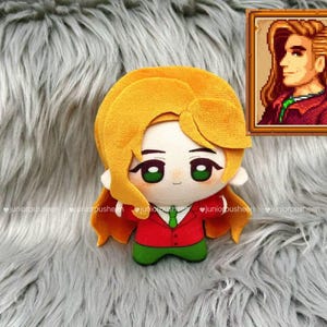 May include: A plush doll of a character with yellow hair, green eyes, and a red jacket with a green tie. The doll is on a grey, fluffy surface. A framed pixel art portrait of the character is in the upper right corner.