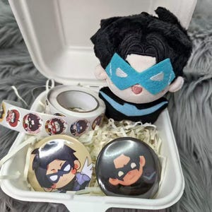 以下が含まれることがあります： 黒い髪と青いマスクのぬいぐるみ、キャラクターのイラストが描かれた円形ステッカーのロール、キャラクターデザインの丸いピンバッジ2つが、白いクラムシェル型の容器に入っています。