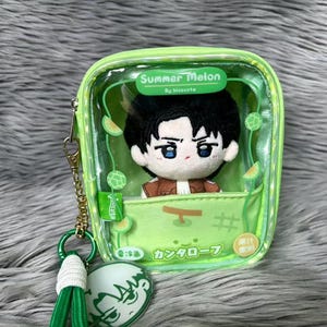 Peut inclure: Une pochette zippée transparente vert citron contenant une poupée en peluche aux cheveux noirs et aux yeux bleus. La pochette porte l'inscription Summer Melon et des caractères japonais sur le devant, avec une chaîne dorée et un cordon vert.