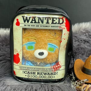 Könnte beinhalten: Ein schwarzer Rucksack mit einer durchsichtigen Vordertasche, in der ein brauner Plüschbär zu sehen ist. Das Einlegeblatt zeigt ein Wanted-Plakat mit roten Blutspritzern, dem Text Wanted und einer Belohnung von 100.000,00 $. Ein kleiner Cowboyhut ist als Anhänger befestigt.