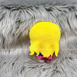 K&ouml;nnte beinhalten: Ein Pl&uuml;schtier mit gelbem Kopf und Haaren, blauen und rosa Akzenten, vor einem grauen, flauschigen Hintergrund. Die gelben Haare des Spielzeugs haben einen gewellten Rand. Der Hintergrund hat eine weiche, pelzige Textur.