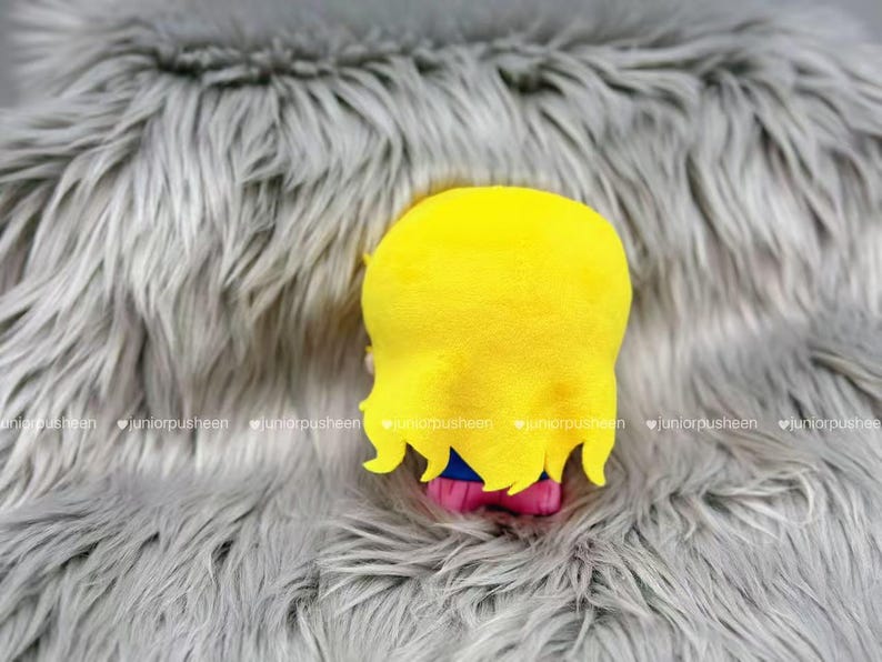 K&ouml;nnte beinhalten: Ein Pl&uuml;schtier mit gelbem Kopf und Haaren, blauen und rosa Akzenten, vor einem grauen, flauschigen Hintergrund. Die gelben Haare des Spielzeugs haben einen gewellten Rand. Der Hintergrund hat eine weiche, pelzige Textur.