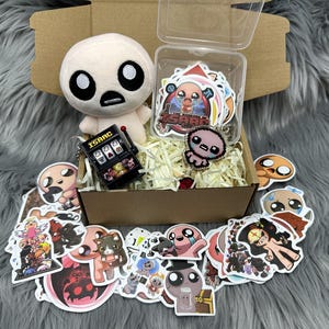 Könnte beinhalten: Eine Geschenkbox gefüllt mit einer Plüschpuppe, Aufklebern und einem Schlüsselanhänger. Die Plüschpuppe hat einen runden Kopf und große Augen. Die Aufkleber zeigen verschiedene Cartoon-Charaktere. Der Schlüsselanhänger ist ein Miniatur-Spielautomat.