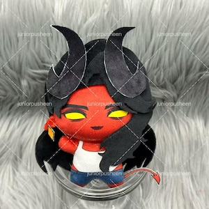 May include: A plush doll with a red face, yellow eyes, and black hair and horns. The doll is wearing a white tank top, blue jeans, and has a red tail. The doll is sitting in a clear glass bowl.