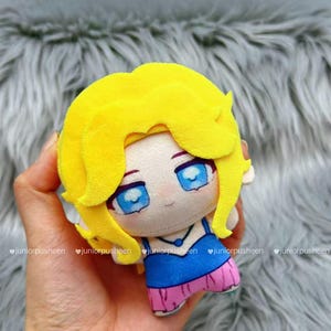 K&ouml;nnte beinhalten: Eine Pl&uuml;schpuppe mit gelben Haaren und blauen Augen. Die Puppe tr&auml;gt ein blaues Oberteil und einen rosa Rock. Die Puppe wird in einer Hand gehalten. Der Hintergrund ist eine weiche, graue, pelzige Textur.