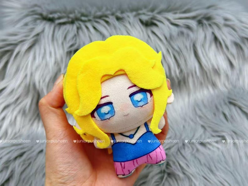 K&ouml;nnte beinhalten: Eine Pl&uuml;schpuppe mit gelben Haaren und blauen Augen. Die Puppe tr&auml;gt ein blaues Oberteil und einen rosa Rock. Die Puppe wird in einer Hand gehalten. Der Hintergrund ist eine weiche, graue, pelzige Textur.