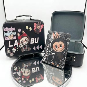 May include: A black rectangular carrying case with a handle and a matching upright rectangular case, both featuring Labubu character graphics, text, and small icons. The set is displayed on a round mirror, showing reflections of the designs.