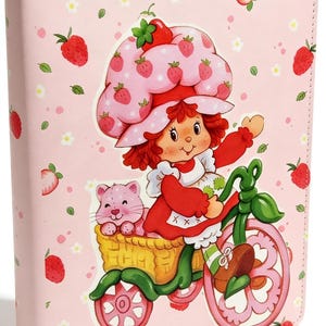 May include: A light pink notebook cover featuring a repeating pattern of red strawberries and small white flowers. The center illustration depicts a character with red hair wearing a strawberry-patterned hat, riding a green tricycle with a pink cat.