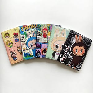 May include: Five rectangular passport holders arranged in a fan shape. Each features a unique graphic print of cartoon characters with rabbit ears, including text like ZIMOMO and LABUBU, in various colors such as peach, blue, green, and black.