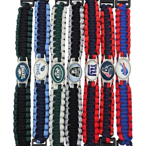 May include: Seven paracord bracelets featuring black plastic side-release buckles and oval metal emblems with various NFL team logos, including the Eagles, Titans, Jets, Raiders, Giants, Texans, and Bills, arranged in a vertical row.