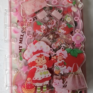 May include: A pink, clear, and decorative binder with Strawberry Shortcake and purse-themed charms. The binder features a variety of pink and red charms, including Strawberry Shortcake characters, strawberries, and miniature purses. The binder is designed with a playful and whimsical aesthetic.