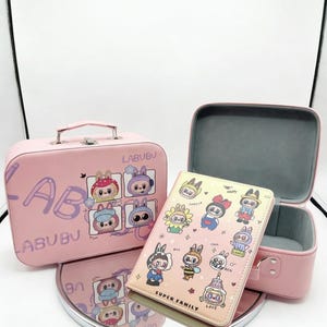 May include: A pink rectangular carrying case with a handle and a matching pink notebook featuring cartoon characters and the text LABUBU and SUPER FAMILY. The case is open, revealing a gray interior, and sits on a round reflective surface.