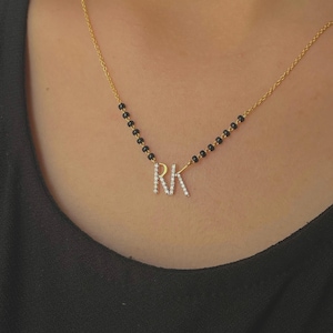 Könnte beinhalten: Eine goldene Halskette mit einer Reihe kleiner schwarzer Perlen an den Seiten und einem zentralen Anhänger in Form der Buchstaben RK, die mit kleinen klaren Steinen besetzt sind. Die Kette wird vor einem schlichten schwarzen Stoffhintergrund gezeigt.
