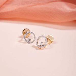 May include: A pair of gold stud earrings featuring a circular frame paved with small white stones and a single centered stone. The earrings have a polished gold backing and a secure post mechanism, showcasing a delicate and refined design.