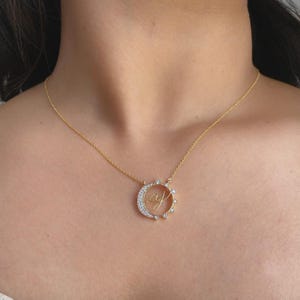 Könnte beinhalten: Eine goldfarbene Halskette mit einem kreisförmigen Anhänger, der auf einer Seite eine Mondsichelform aufweist, die mit kleinen klaren Steinen besetzt ist. In der Mitte des Anhängers befinden sich die kursiven Buchstaben aj in Gold.
