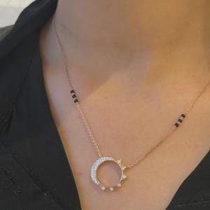以下が含まれることがあります： 外縁に小さな透明な石があしらわれた円形のペンダントが付いたゴールドトーンのネックレス。繊細なチェーンには、中央のペンダントの両側に左右対称に配置された3つの小さな黒いビーズのセグメントが2つ含まれています。