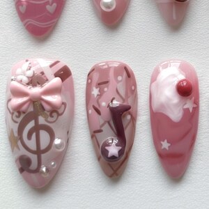 May include: A collection of pink and white nail art designs. One nail features a treble clef and bow, another a musical note, and another a cherry on top of a white swirl. Other nails have hearts and stars.