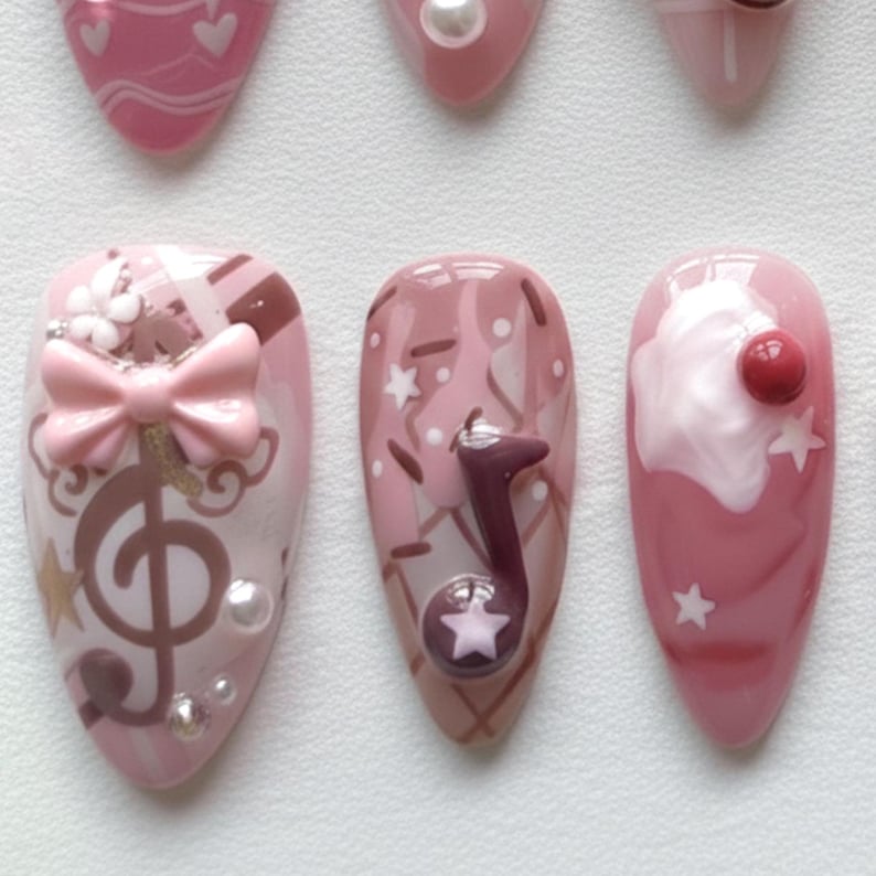 May include: A collection of pink and white nail art designs. One nail features a treble clef and bow, another a musical note, and another a cherry on top of a white swirl. Other nails have hearts and stars.